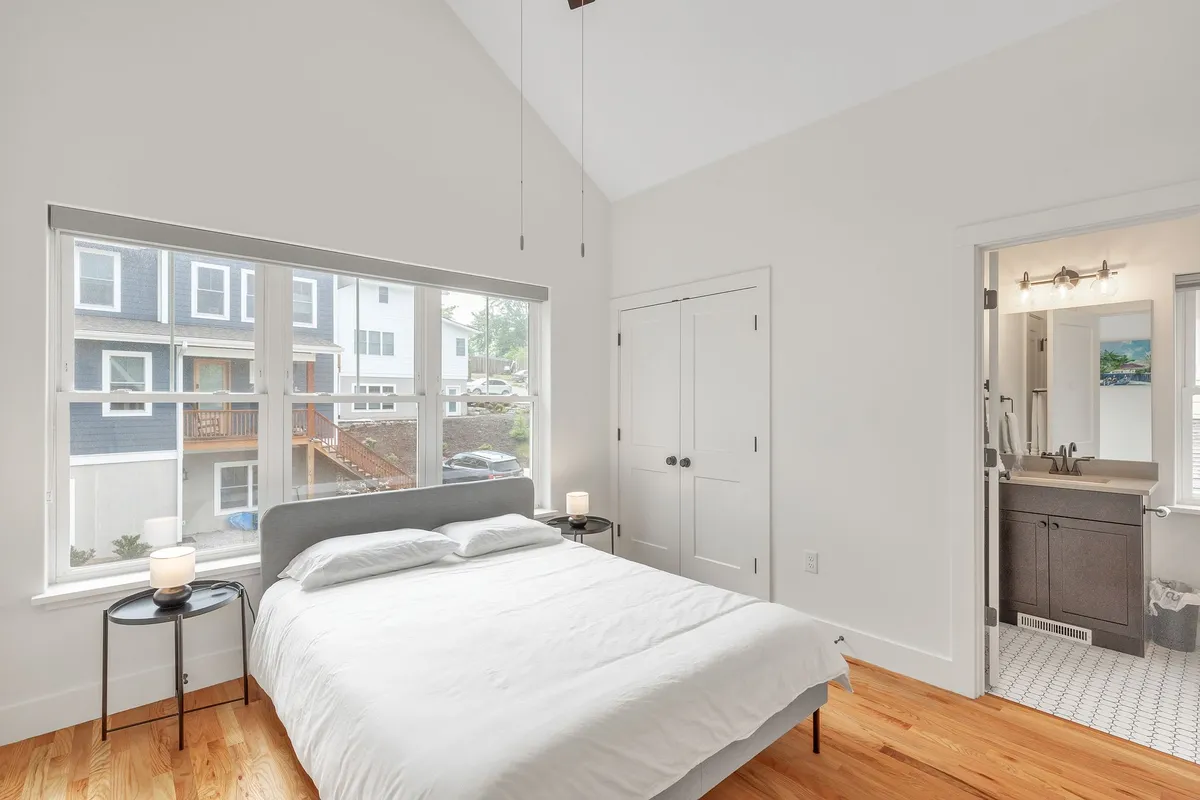 This image features a serene bedroom with a bed dressed in white bedding, large windows allowing in natural light, a bathroom doorway nearby, and a nightstand with a lamp. The peaceful setup and soft light create a restful atmosphere ideal for relaxation.