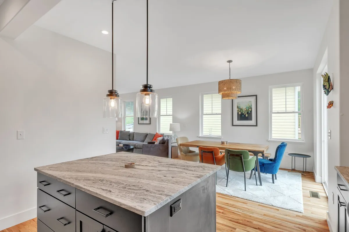 A wide view of the open-plan living area, featuring a kitchen island, pendant lights, a cozy dining area, and a plush sofa. Expansive windows allow natural light to flood the space, and the layout encourages easy movement between relaxation and dining zones.