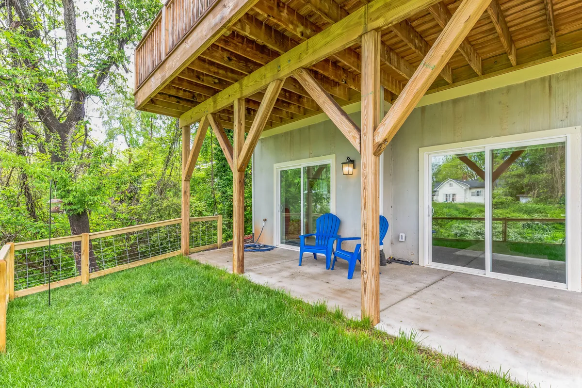 An exterior view of a patio area with blue chairs, wooden beams, green grass, and sliding glass doors that connect directly to the interior. The setup invites relaxation while offering a visual connection between indoor and outdoor spaces overlooking greenery.
