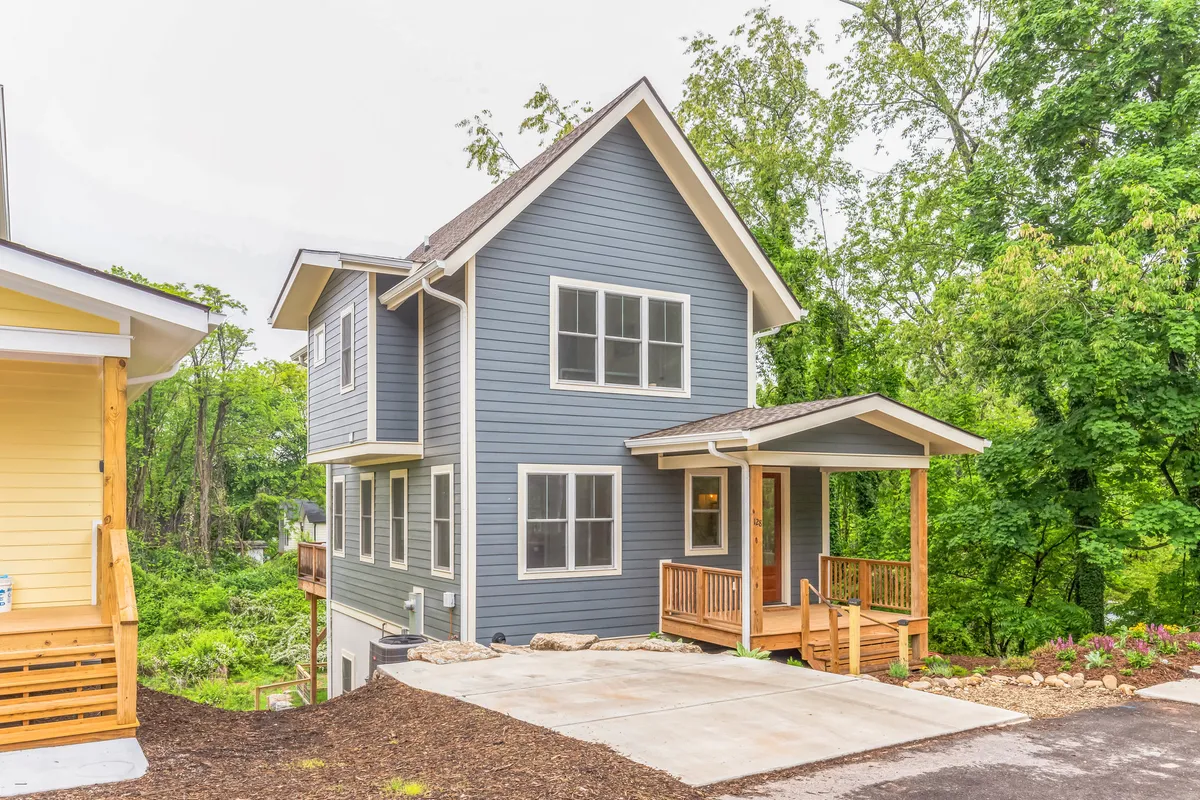 The image features the exterior of a newly constructed single-family home with striking blue siding and a spacious wooden porch, surrounded by lush greenery. The setting feels peaceful and inviting, with ample space for outdoor gatherings or relaxing while enjoying nature. The porch offers an ideal spot for unwinding at sunset or spending quality time with family and friends.