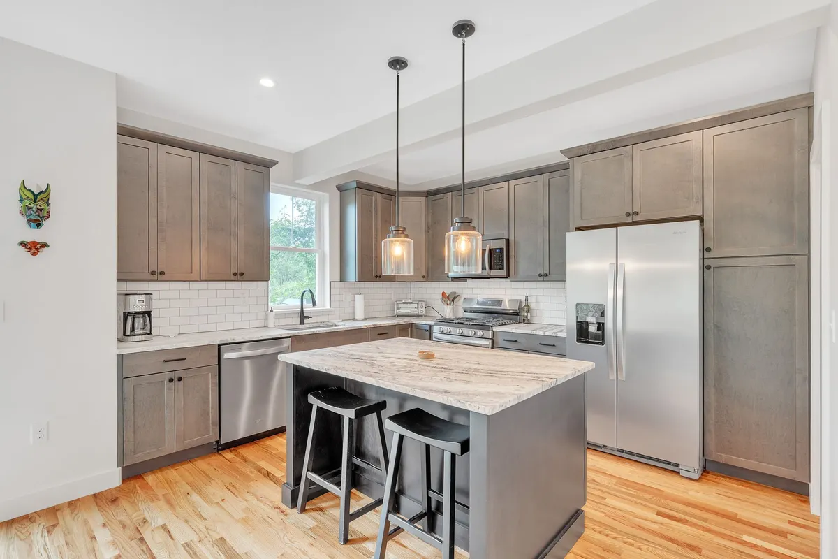 A warm, contemporary kitchen featuring a spacious central island with pendant lights overhead, sleek stainless steel appliances, and wooden flooring. The open layout hints at easy movement between cooking, dining, and living spaces.