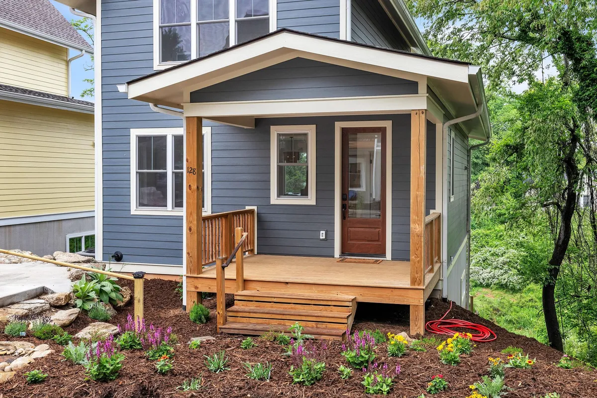The image features a charming front porch with flower beds, wooden steps, and an inviting entrance door. The space is set up for morning coffee, meeting guests, or simply enjoying the outdoors, reinforcing the sense of community and easy access to neighborhood activities.