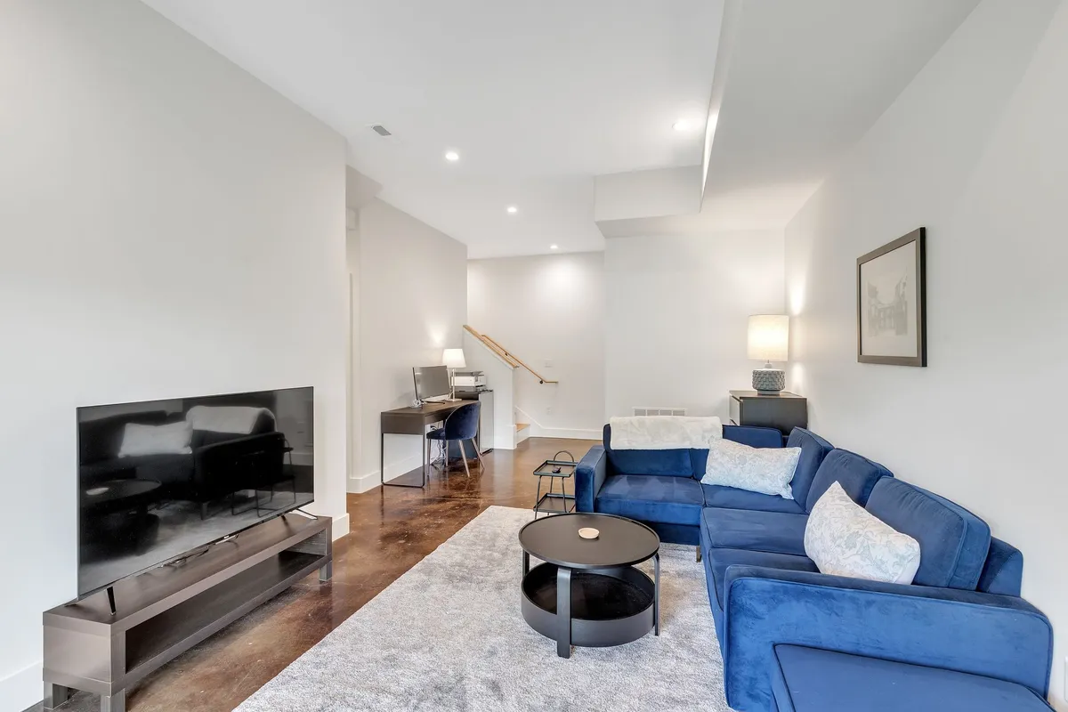 A modern, cozy lower-level room featuring a blue sectional sofa, black coffee table, TV stand, and, notably, a dedicated desk with chair adjacent to a staircase. The workspace includes a monitor and is situated to provide a quiet, productive environment while remaining integrated with the comfortable family space.